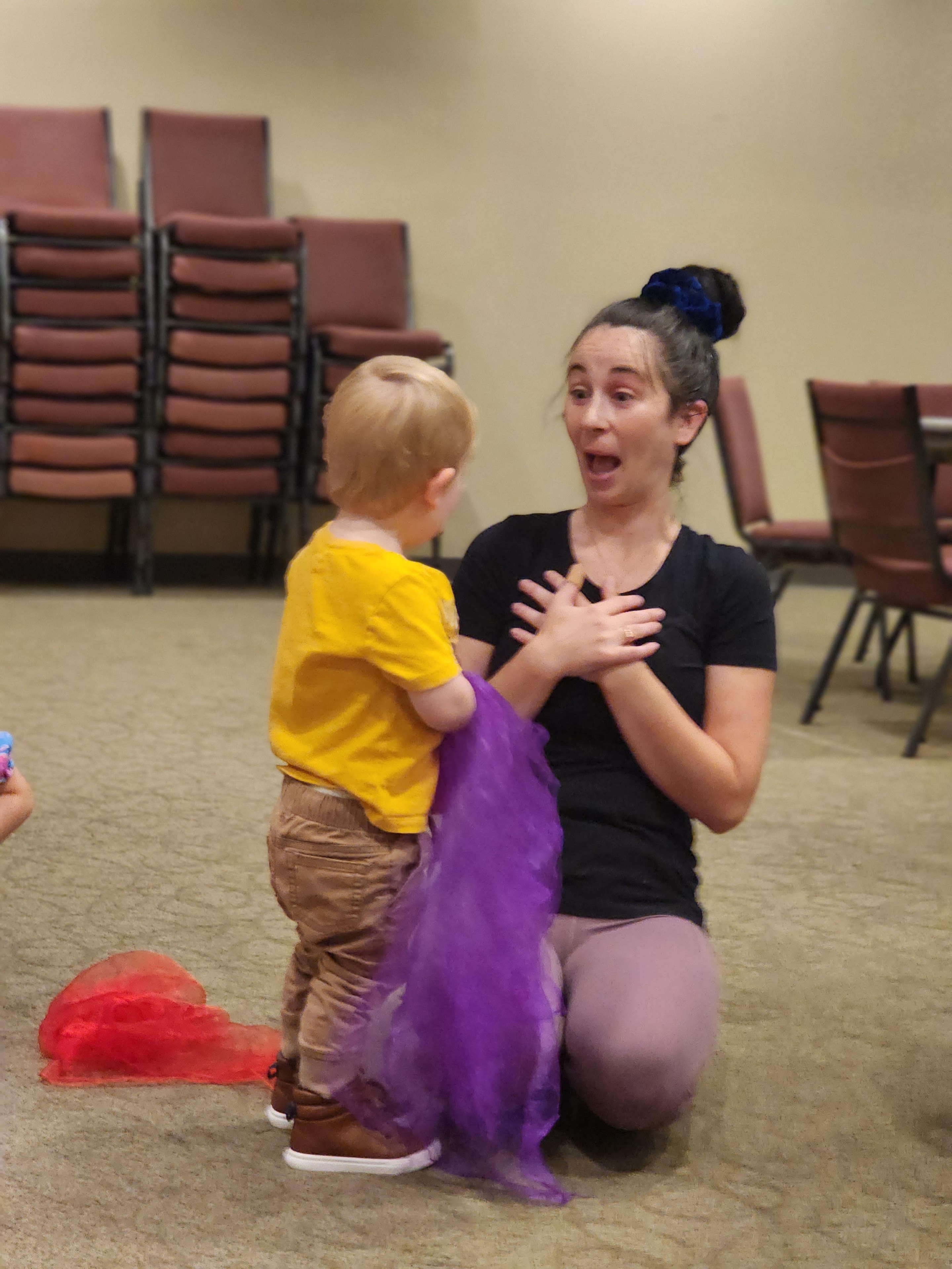 A mom kneeling with a surprised expression as her toddler holds a purple scarf during Zumbini