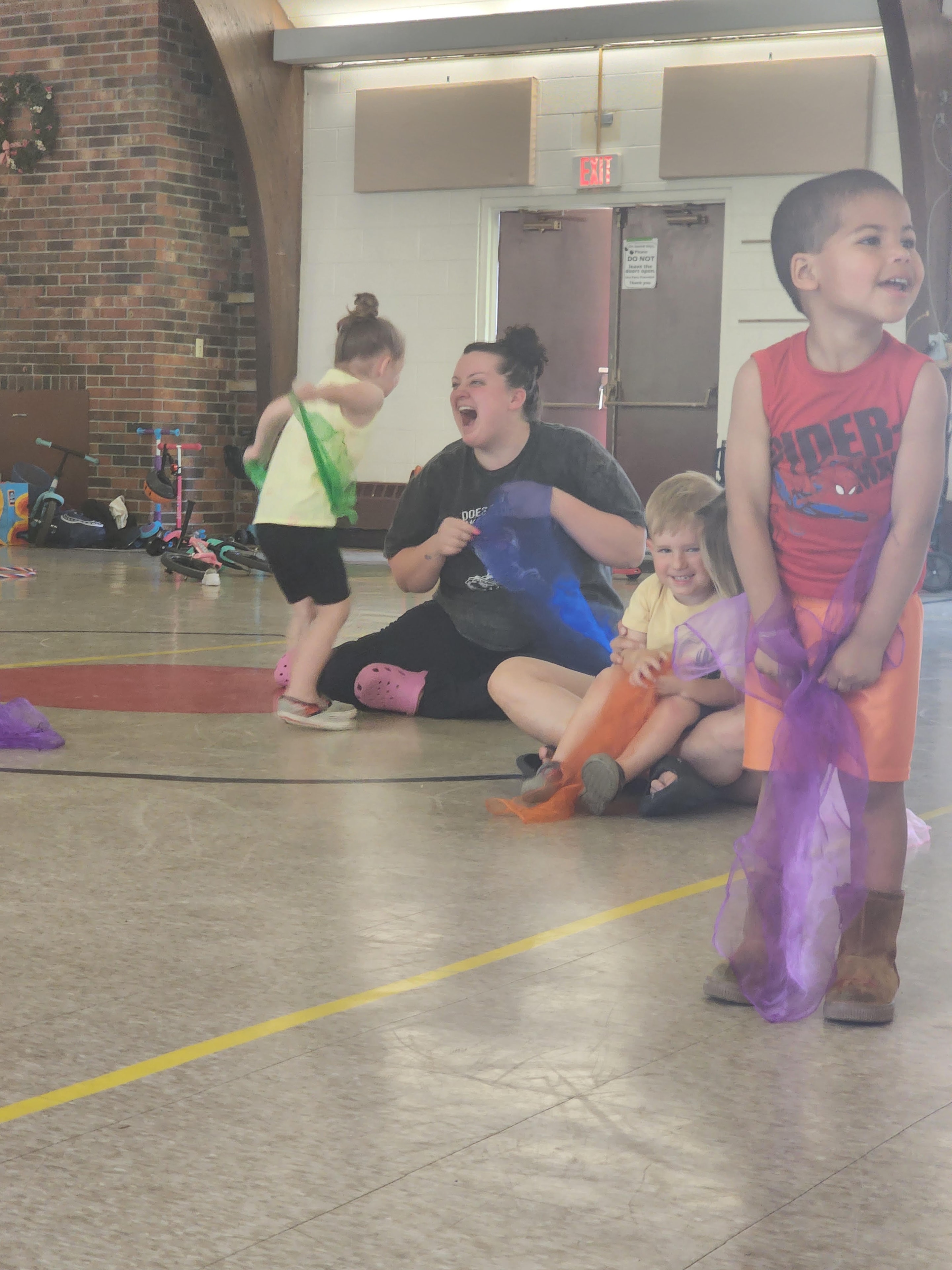 A mom laughing with kids waving colorful scarves during a Zumbini class in a gymnasium