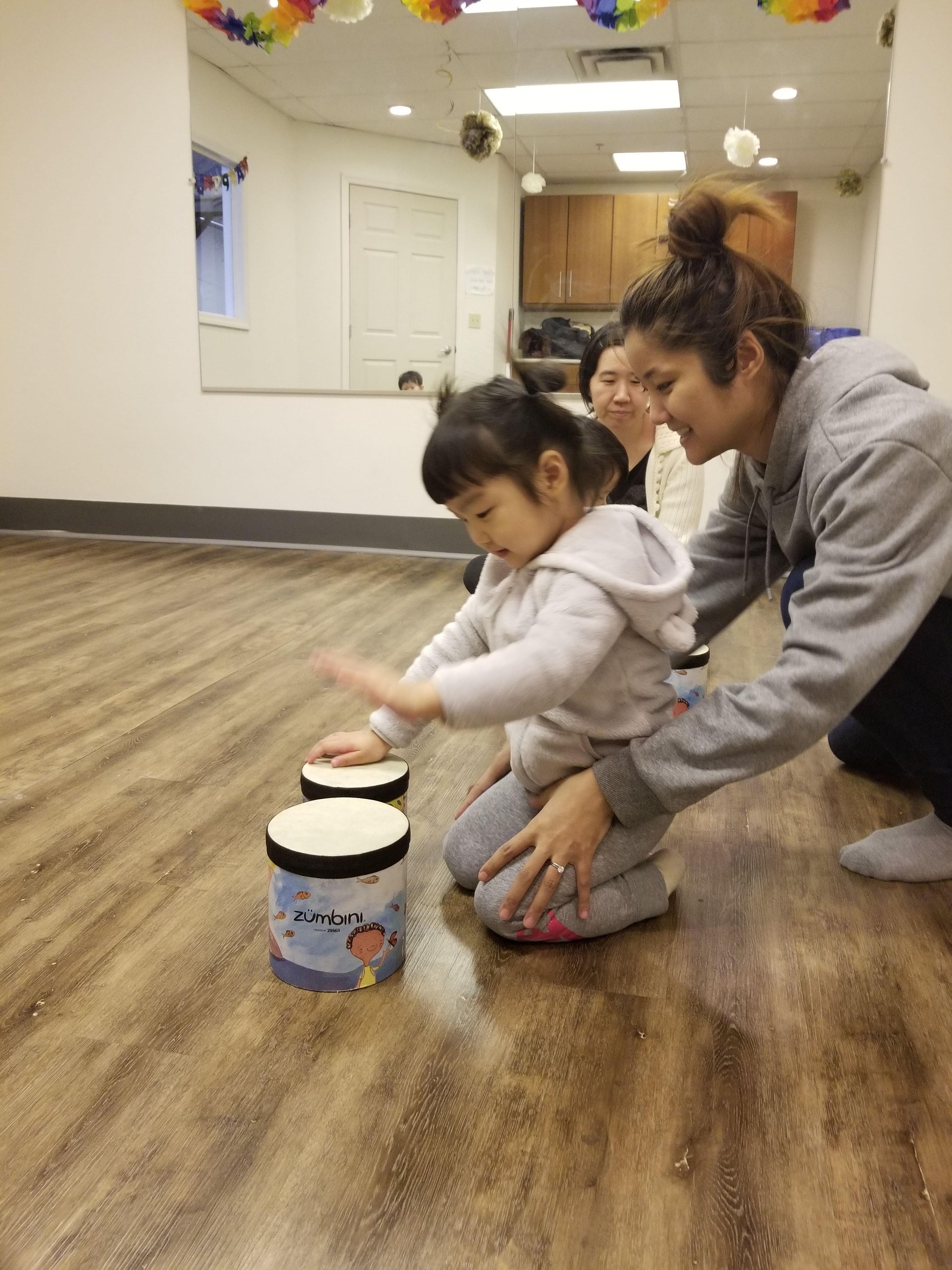 A mom helping her toddler play the Zumbini drum on the floor during class